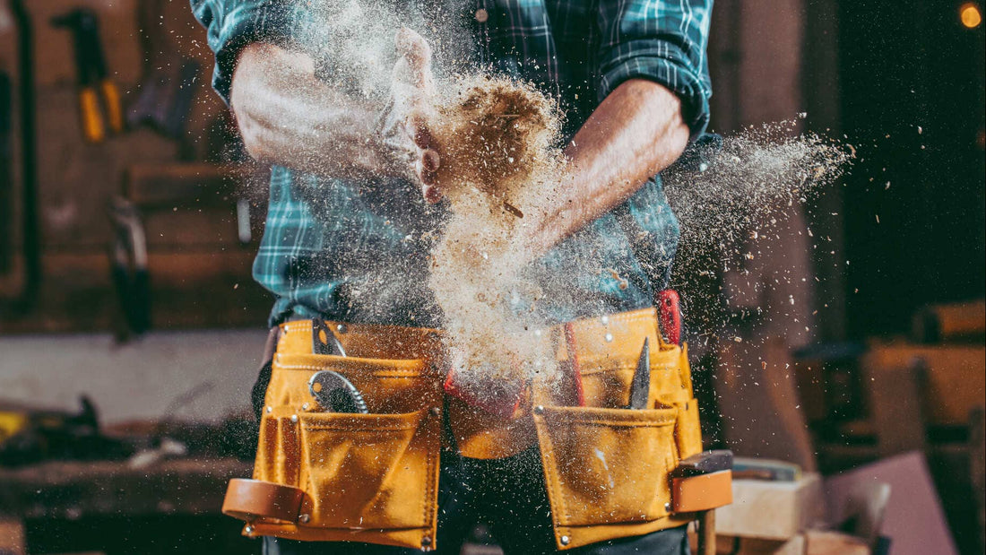 Person in a workshop with tools and wood shavings, wearing a yellow tool belt.